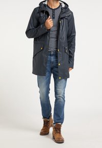 Veste imperméable marine avec capuchon, boutons-pression jaunes et deux poches avant, portée par-dessus une chemise à manches longues rayée et un jean bleu.