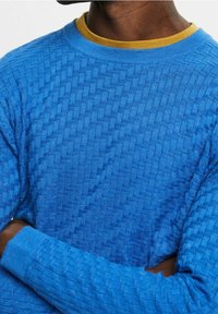 Blauer strukturierter Pullover mit einem gewebten Muster, Rundhalsausschnitt und gerippten Ärmeln, über einem gelben Innenhemd getragen.