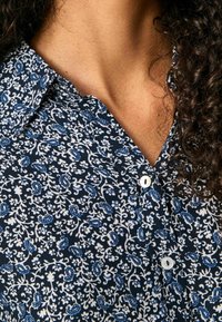 Nærbillede af en persons hals og krøllet hår iført en marineblå bluse med hvide og blå blomster mønstre og hvide knapper.