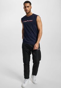 Marineblå ermeløs t-skjorte med rød tekstlogo, kombinert med svarte bukser med lommer og hvite sneakers, som skaper et kontrasterende antrekk.