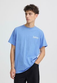 T-shirt en coton bleu clair à coupe décontractée, col rond, avec le texte blanc "Soulscape" sur la poitrine gauche. Porté avec un pantalon noir.