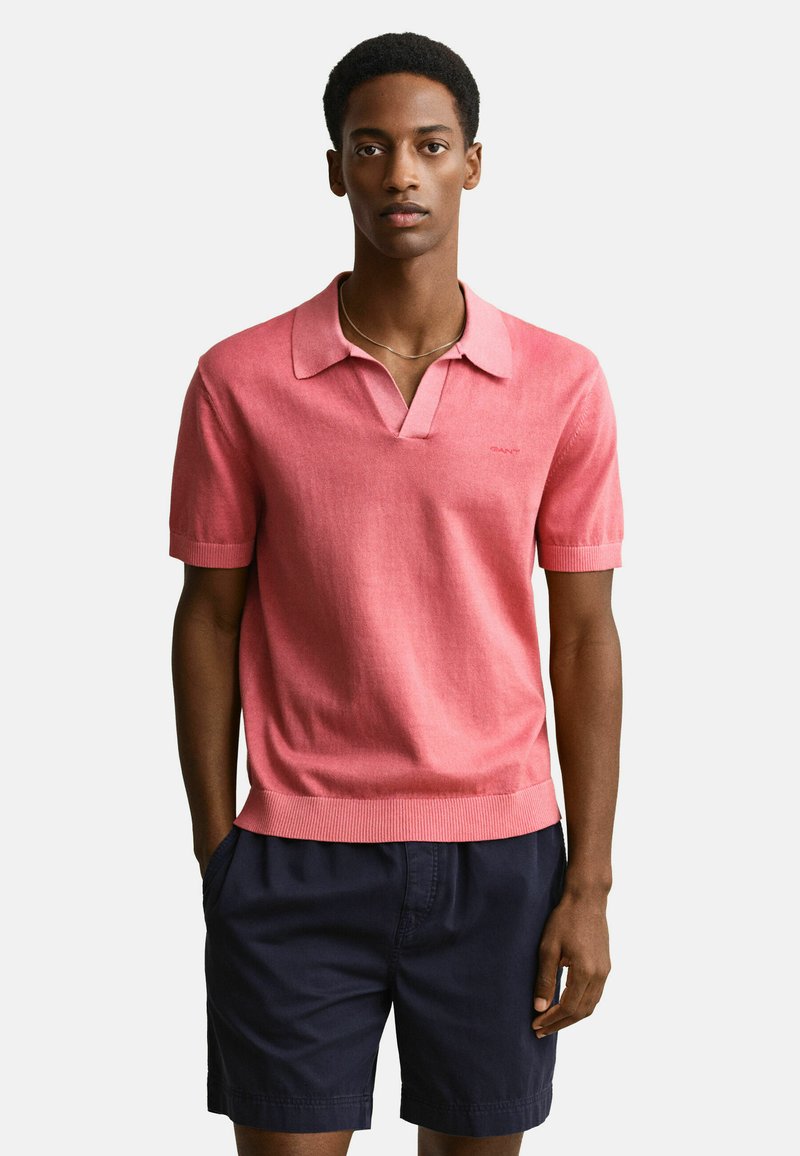 Jeune homme portant une chemise corail à manches courtes avec col et un short bleu marine foncé, debout les mains dans les poches devant un fond uni.