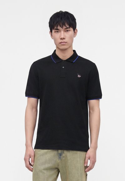 Jeune homme portant un polo noir avec une bordure bleue et un pantalon vert clair, se tenant devant un fond blanc uni.