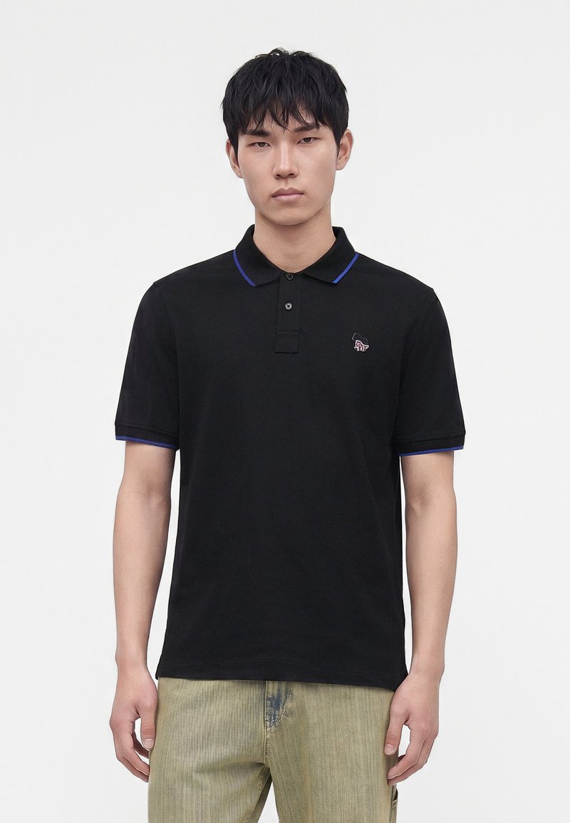 Jeune homme portant un polo noir avec une bordure bleue et un pantalon vert clair, se tenant devant un fond blanc uni.