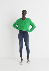 Vihreä, pörröinen oversized-neule V-kauluksella, yhdistettynä tummansinisiin tiukkoihin farkkuihin ja valkoisiin urheilukenkiin. Yksinkertainen tausta.