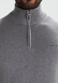 Gros plan d'un homme portant un pull en maille grise côtelée avec un col à demi-fermeture éclair, montrant une partie de sa barbe et le nom de la marque sur la poitrine.