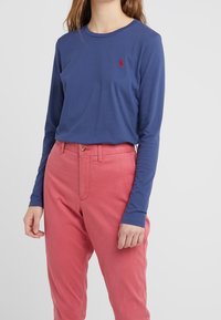 Dlouhý rukáv košile v námořnické bavlně s kulatým výstřihem a červeným logem na hrudi, spárované s růžovými bavlněnými kalhotami s vysokým pasem.