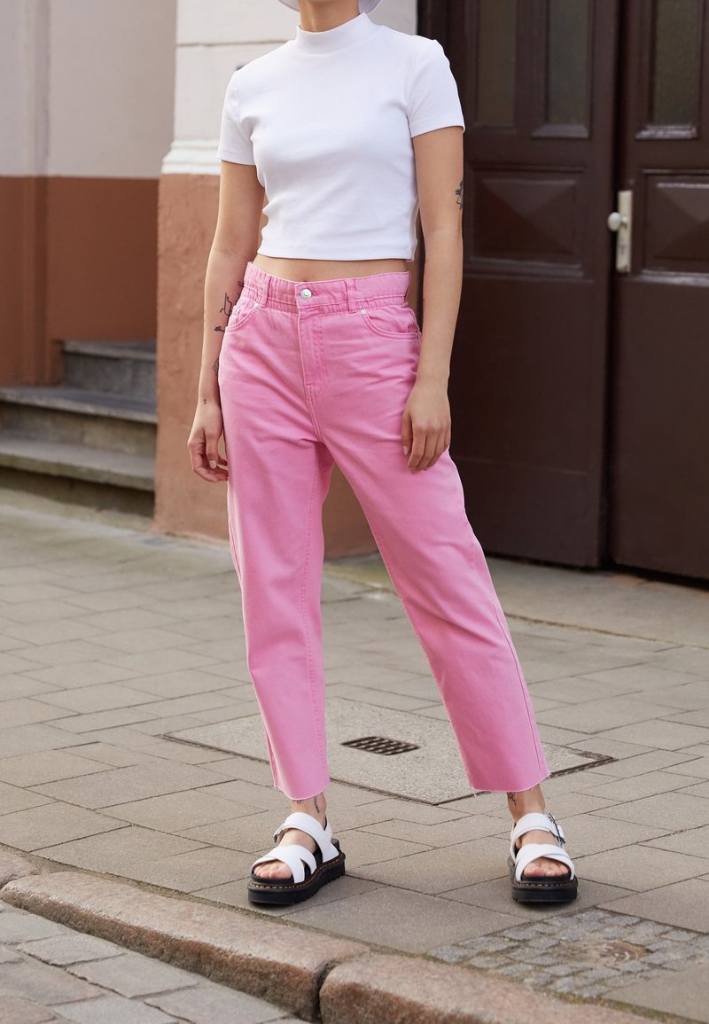 Personne portant un col roulé court blanc, un jean rose taille haute, des sandales blanches, debout sur un trottoir carrelé près d'une porte marron.