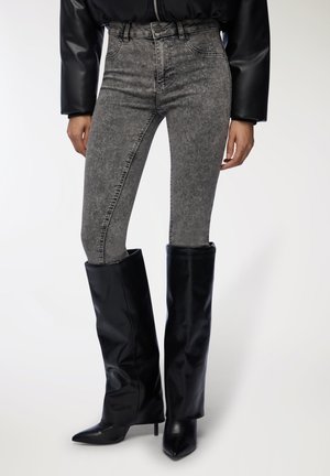 I jeans a vita alta grigi sbiaditi presentano una vestibilità skinny, abbinati a stivali neri lucidi sopra il ginocchio con tacco a punta.