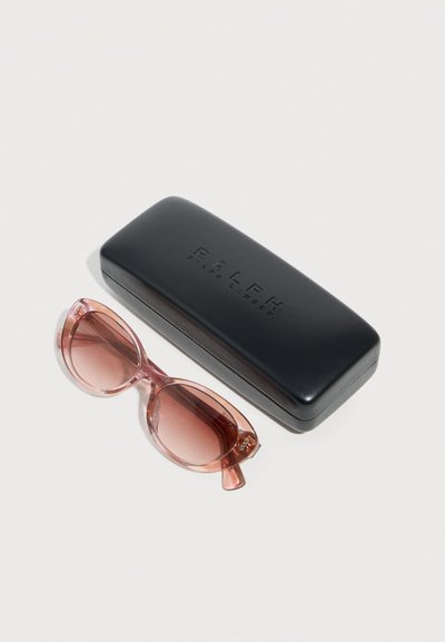 Στρογγυλά γυαλιά ηλίου με ροζ απόχρωση δίπλα σε κλειστή μαύρη σκληρή θήκη με ανάγλυφη τη φράση "RALPH Ralph Lauren" πάνω σε λευκή επιφάνεια.