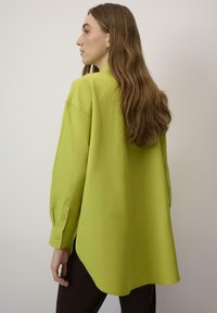 Camicia button-up oversize verde lime con maniche lunghe, orlo curvo e colletto; realizzata in tessuto leggero; abbinata a pantaloni scuri.