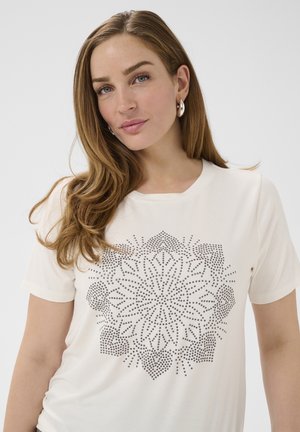 Junge Frau mit langen hellbraunen Haaren, die silberne Creolen und ein weißes T-Shirt mit einem schwarz gepunkteten, floralen Mandala-Design trägt.
