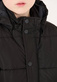 Giacca a vento nera con colletto alto, texture trapuntata e bottoni a pressione neri, progettata per calore e comfort.