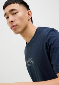 Tricou albastru marin din bumbac, cu guler rotund și mâneci scurte; are un text brodat alb și galben pe piept.