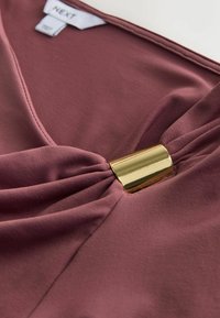 Mauve Stoffoberteil mit V-Ausschnitt, ausgestattet mit einer Faltendetail in der Mitte und einem goldenen Metallakzent. Glatte Textur, saubere Nähte.