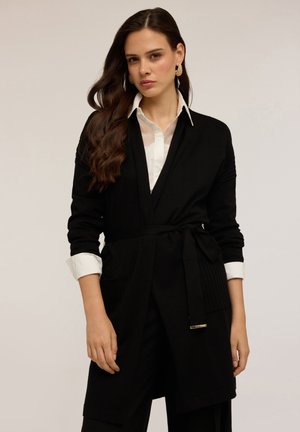 Cardigan noir avec une ceinture à nouer, manches longues et détails côtelés. Porté par-dessus une chemise blanche, mettant en valeur un look élégant. Texture lisse.