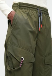 Žali krovinių kelnių modelis su elastinga juosta, šoninėmis kišenėmis ir didelėmis krovinių kišenėmis. Yra rožinė sagtis ir dekoratyviniai pilki raiščiai.