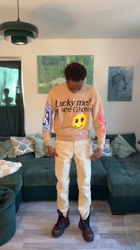 Személy áll a nappaliban, bézs pulóvert visel "Szerencsés vagyok! Látok szellemeket" felirattal és mosolygós arccal, krémszínű nadrágot és sötét csizmát.