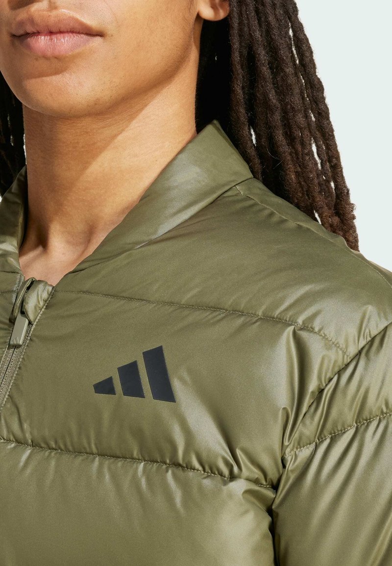 Una persona con largas rastas oscuras lleva una chaqueta de plumas verde con cremallera que presenta un logotipo negro en la zona del pecho.