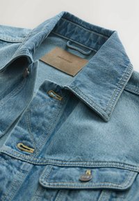 Világoskék farmerdzseki hajtogatott gallérral, begombolt zsebfedővel és látható varrással egy fehér felületen.