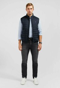Gilet matelassé marine avec col montant, fermeture zip, porté sur une chemise bleu clair. Associé à un jean foncé et des baskets blanches. Design minimaliste.