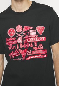 Schwarzes T-Shirt mit kurzen Ärmeln, das verschiedene rote und weiße Jordan-Logos und Designs auf der Vorderseite zeigt, einschließlich grafischem Text und Formen.