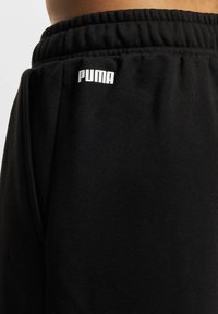 Svarta mjukisbyxor gjorda av mjukt tyg med elastisk midja. Har en framträdande vit Puma-logotyp på vänster sida. Enkelt design.