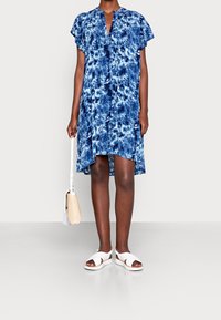 Modrá batikovaná šaty s krátkými volánovými rukávy, lehký materiál, tvar do A a výstřih s kulatým otvorem. Vybavené bílými sandály a kabelkou.