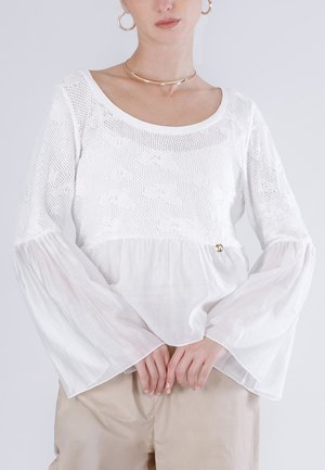 Femme portant un chemisier blanc à manches longues avec un haut en maille florale texturée et des manches évasées transparentes, un pantalon beige, des boucles d'oreilles créoles dorées et un collier ras du cou.