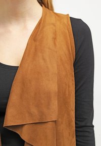 Chaleco de ante marrón con diseño delantero drapeado, usado sobre una blusa negra, con una textura suave y sin cierres o adornos visibles.