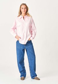 Kvinde iført en løs lyserød skjorte med knapper, blå vide jeans og taupe balletflade sko, stående med hænderne i lommen.