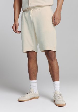 Beige katoenen shorts met een elastische tailleband en een trekkoord. Soepele textuur, knielang, gecombineerd met witte sokken en beige sneakers.