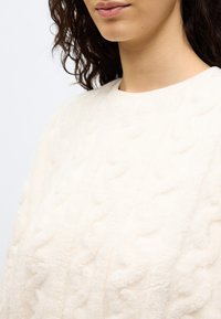 Γυναίκα που φοράει ένα απαλό, εκρού πουλόβερ πλεκτό με σχέδιο κοτσίδα και στρογγυλή λαιμόκοψη, εμφανιζόμενο από τον λαιμό έως  το μέσο του κορμού.