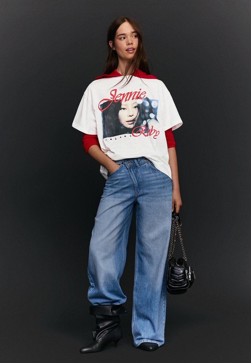 Junge Frau trägt ein weißes T-Shirt mit der Aufschrift "Jennie Ruby" über einem roten Langarmshirt, dazu lockere blaue Jeans und schwarze Stiefel, hält eine schwarze Handtasche mit Kettenriemen.