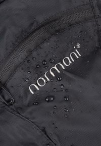 Schwarzer wasserdichter Stoff mit sichtbaren Wassertropfen, ausgestattet mit einer Reißverschlusstasche und dem silbernen Markenlogo "normani" gut sichtbar angebracht.