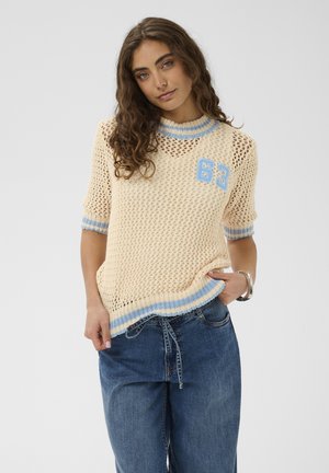 Jeune femme aux cheveux bouclés portant un haut en maille beige avec des bordures bleues et le numéro 63, associée à un jean bleu ample et un bracelet argenté.