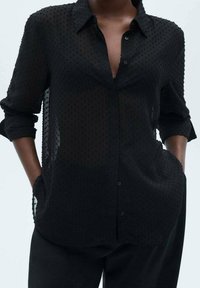 Camicia nera trasparente con pattern testurizzato, maniche lunghe, colletto e tasche ai lati. Progettata per una vestibilità ampia.