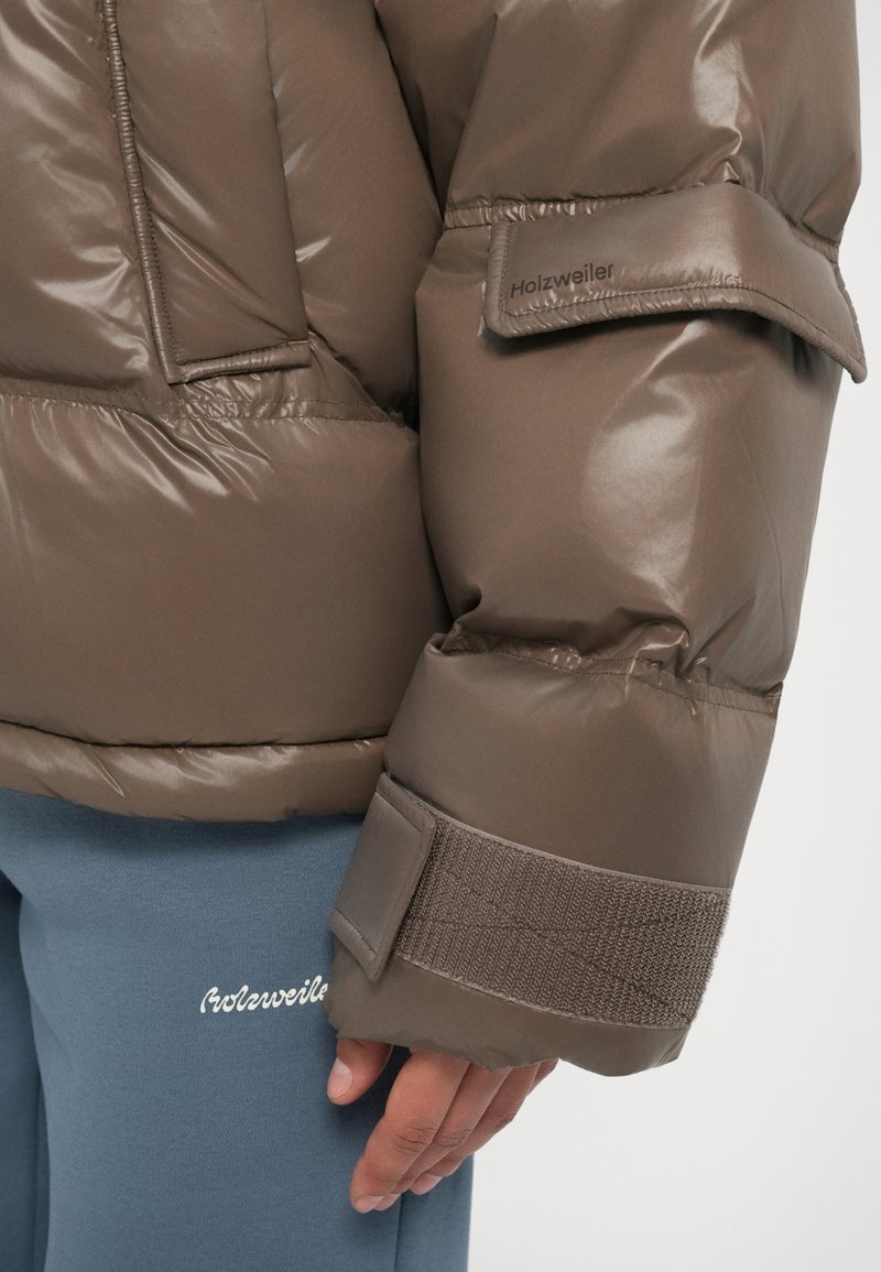 Close-up van een persoon die een bruine gewatteerde jas met klittenbandriemen en blauwe broek met geborduurd logo op de dij draagt.