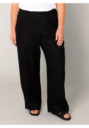 Volwassene draagt losse zwarte broek, witte top en zwarte sandalen, staand tegen een effen lichte achtergrond.