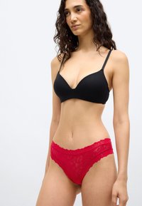 Γυναίκα με σγουρά μαλλιά, φορώντας μαύρο σουτιέν και κόκκινο δαντελένιο εσώρουχο, στέκεται μπροστά σε έναν απλό ανοιχτόχρωμο φόντο.