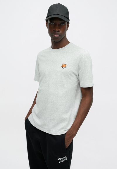 Tricou gri cu maneci scurte, cu un broderie a unei cap de vulpe portocalie pe piept, purtat cu pantaloni negri care au detalii din text alb pe coapsă.