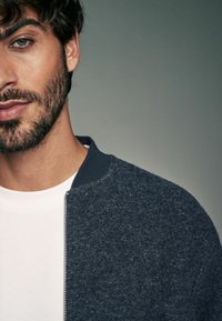 Marineblauwe wollen bomberjack met een gestructureerde afwerking, voorzien van een ritssluiting en een contrasterende donkere kraag, gedragen over een wit T-shirt.
