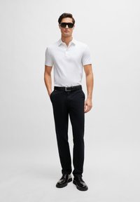 White short-sleeve polo shirt paired with black trousers and a black belt. Model wears dark sunglasses and black shoes, standing against a neutral background.