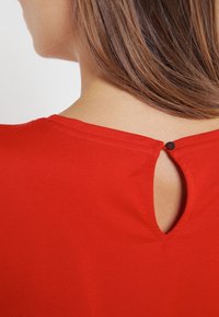 Czerwona koszulka z zaokrąglonym dekoltem i małym wycięciem w kształcie klucza z tyłu, zapięta czarnym guzikiem. Gładki, elastyczny materiał.