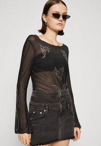 Svart genomskinlig bodysuit med långa ärmar som har dekorativt silverbroderi och utsvängda ärmslut, tillsammans med en hög midja svart denim minikjol.