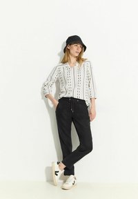 Pantaloni neri con cordino abbinati a una blusa bianca a righe. La modella indossa un cappello a secchiello nero e sneakers bianche con dettagli neri.