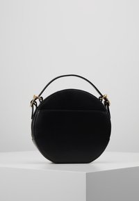 Kulatá černá kožená kabelka s horním uchem a zlatými sponami, vystavená na bílém geometrickém podstavci na jednobarevném šedém pozadí.