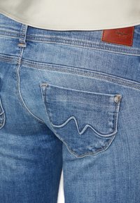 Modré džínové kalhoty s vlnitým vzorem na zadní kapse, se kontrastním prošíváním a hnědou koženou nášivkou značky.