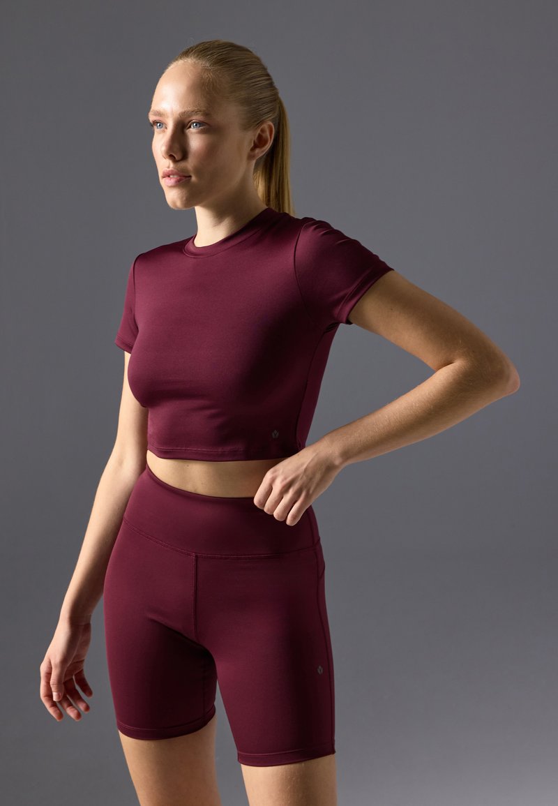 Maroon atletski crop top s kratkim rukavima i odgovarajućim visokim biciklističkim šortsevima, izrađen od glatkog, rastezljivog materijala. Jednostavan dizajn i kroj.