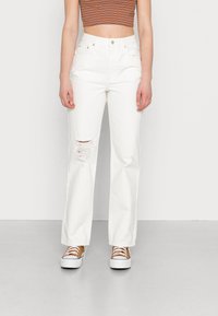 Jeans brancos de perna reta com detalhes desgastados no joelho, combinados com ténis castanhos e brancos. A parte de cima é uma t-shirt cropped de riscas multicoloridas.